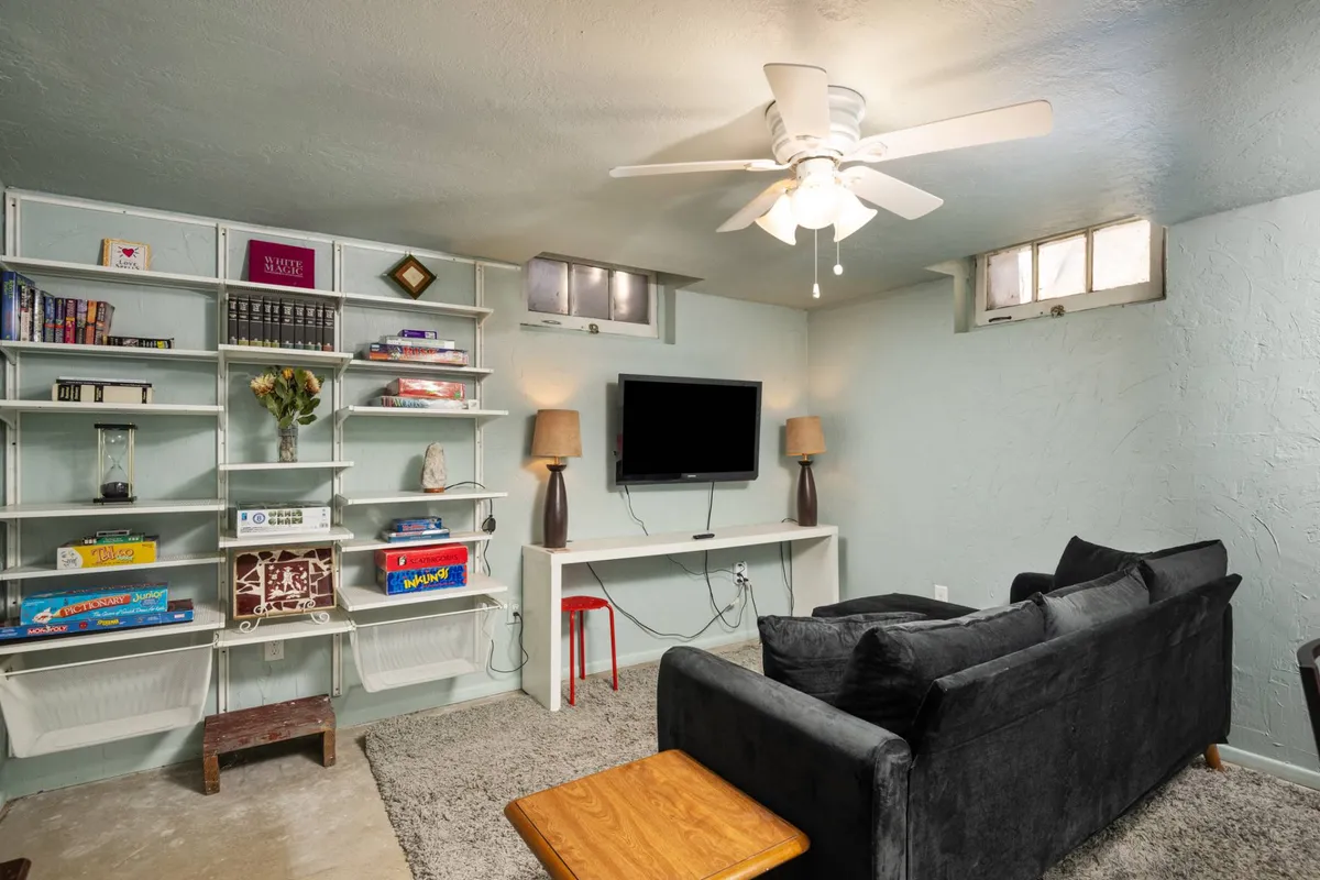 A basement living area with shelves brimming with books and games, a plush sofa, a television, and softly glowing lamps. The setting suggests a welcoming space for reading, movie nights, or gathering with close friends.