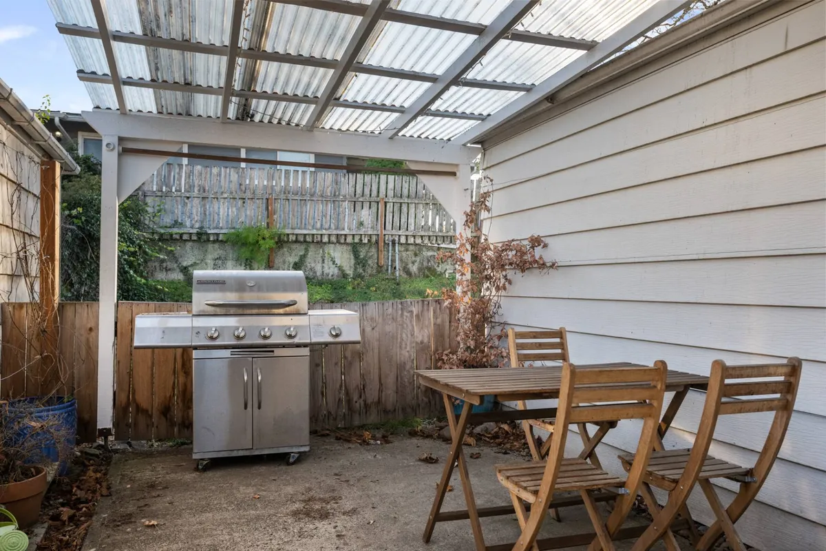 An inviting backyard setup, complete with a grill, table and chairs under an overhead cover, and a fenced-in area. The scene is arranged for both casual barbecues and relaxed outdoor meals, hinting at lively social gatherings and family moments in the fresh air.