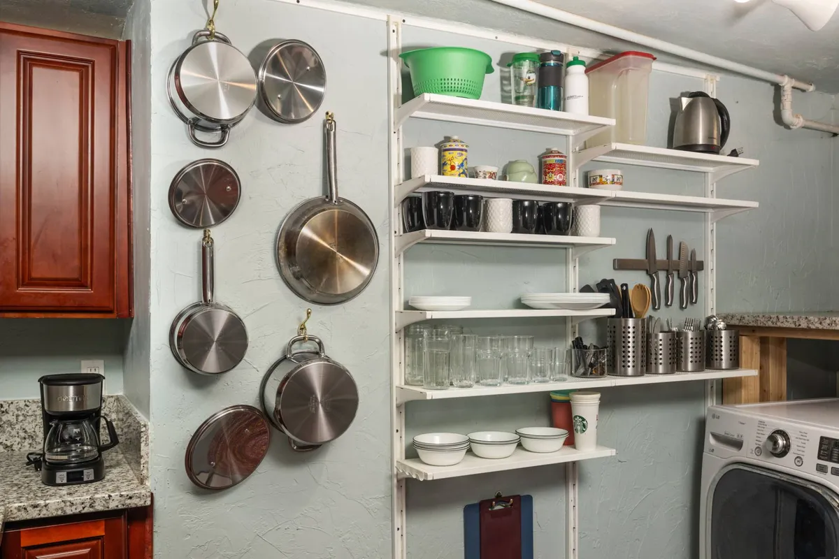 A tidy, homey kitchenette corner featuring metal cookware, shelves neatly organized with dishes, and a coffee maker set on the counter. The space expresses readiness for meal prep and casual morning rituals.