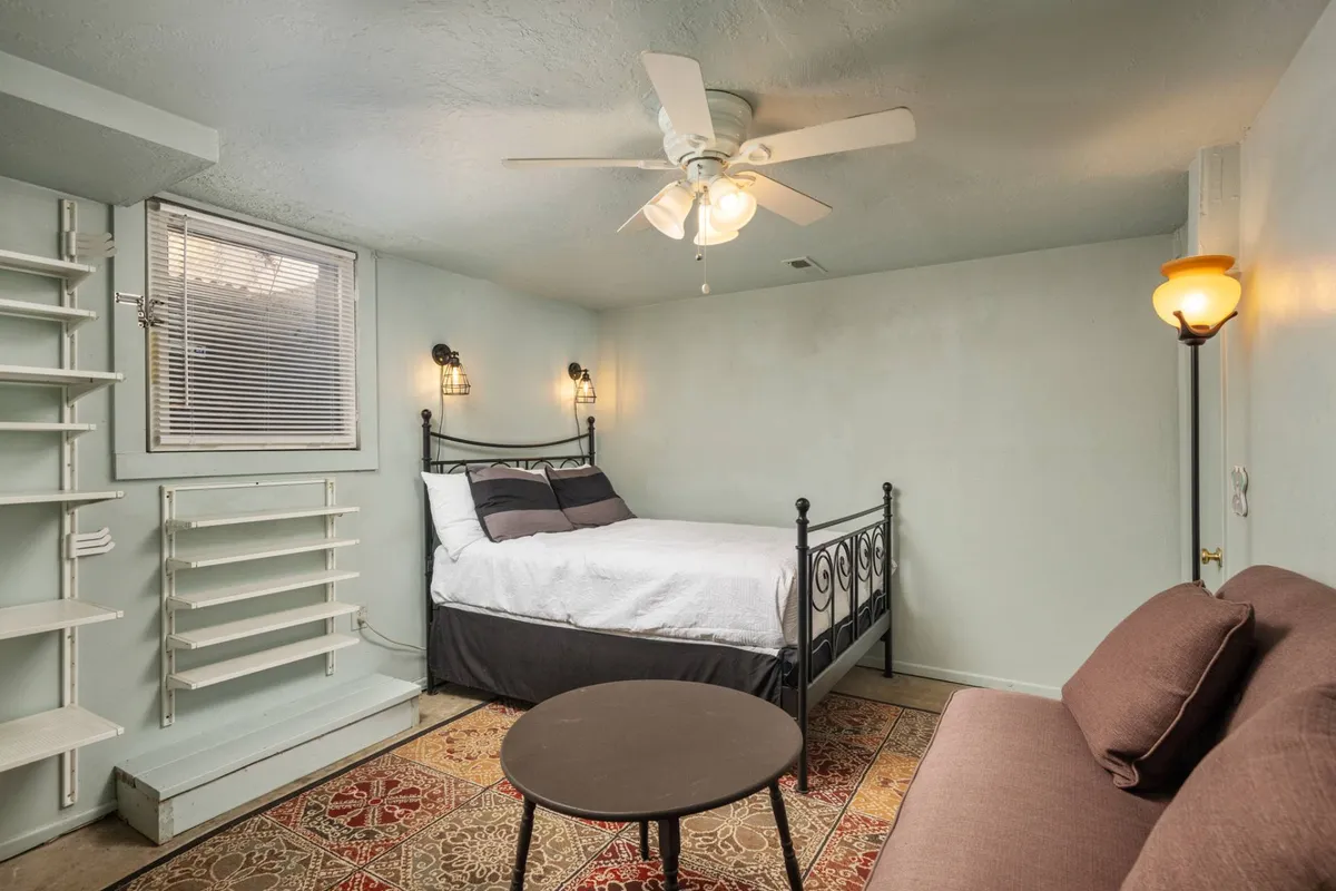 A welcoming bedroom scene with a comfortable bed, a plush rug, a cozy sofa, and open shelving. Natural light filters gently through the window, enhancing the room's inviting atmosphere.