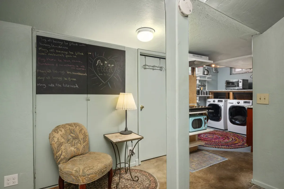 A basement hallway scene featuring a chalkboard with handwritten messages, a comfortable chair placed on a decorative rug, and a view of the laundry area. The lighting is soft, creating a cozy and welcoming atmosphere ideal for relaxation and personal moments.