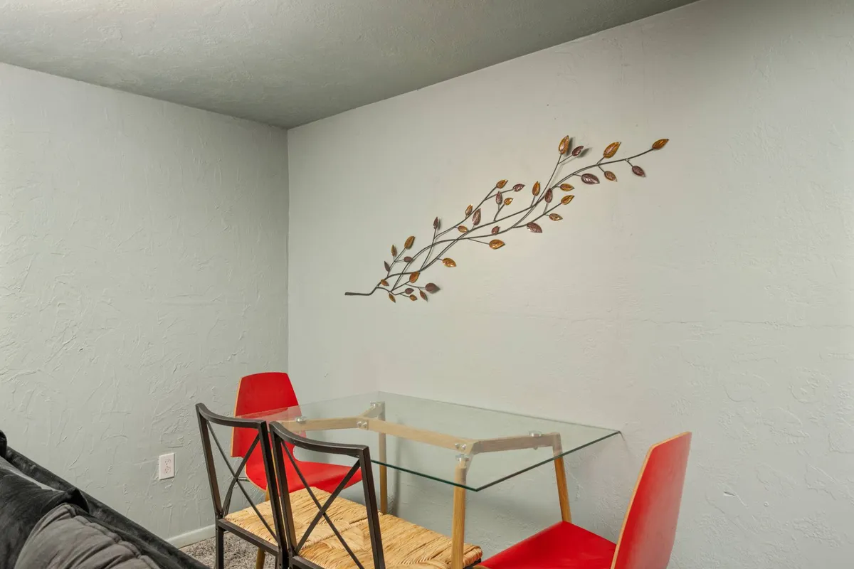 A bright, contemporary dining area featuring a glass table surrounded by vibrant red chairs and metal wall art. The ambiance suggests a space where guests can enjoy a meal, get some work done, or socialize in comfort before or after their travels.