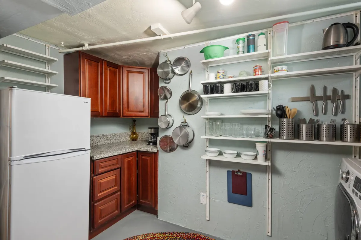 This image showcases a compact interior kitchen space featuring wooden cabinetry, a granite countertop, open shelving stocked with kitchenware, and essential kitchen appliances. The layout conveys practicality and warmth, ideal for quick meal preparations or brewing morning coffee.